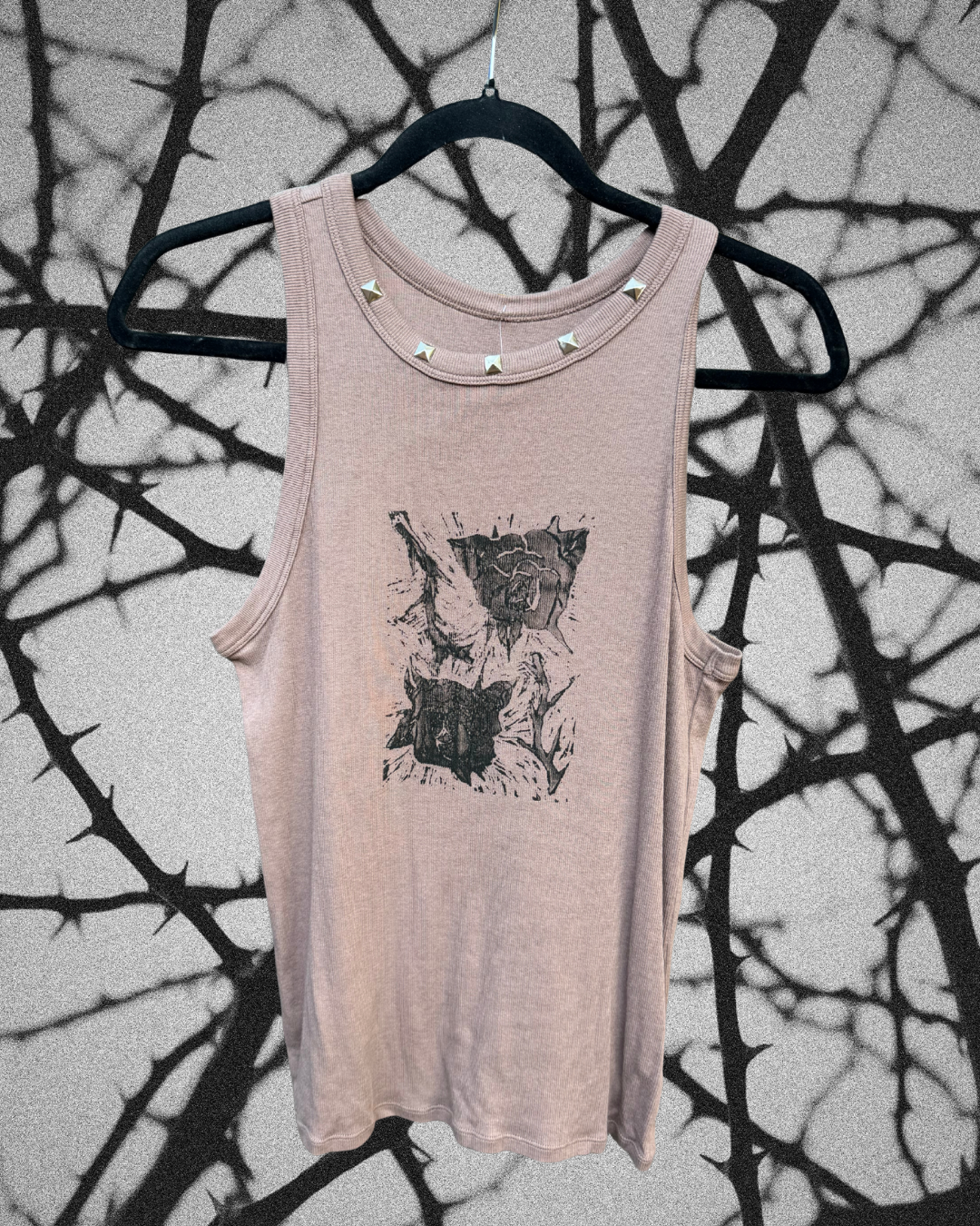 A sleeveless light pink tank top with a black graphic rose print on the front and silver studs along the neckline, hanging on a black hanger against a wall with tree branch shadows.