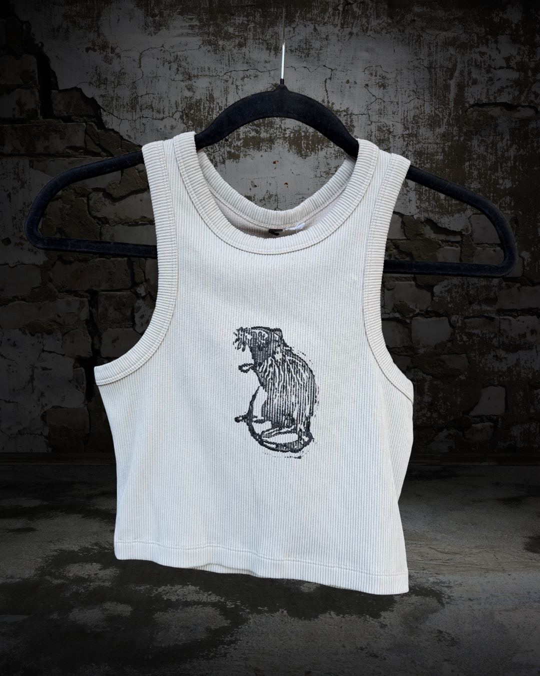 A ribbed tan sleeveless tank top with a black line art graphic of a rat on the front, hanging on a black hanger against a dark, textured brick wall.