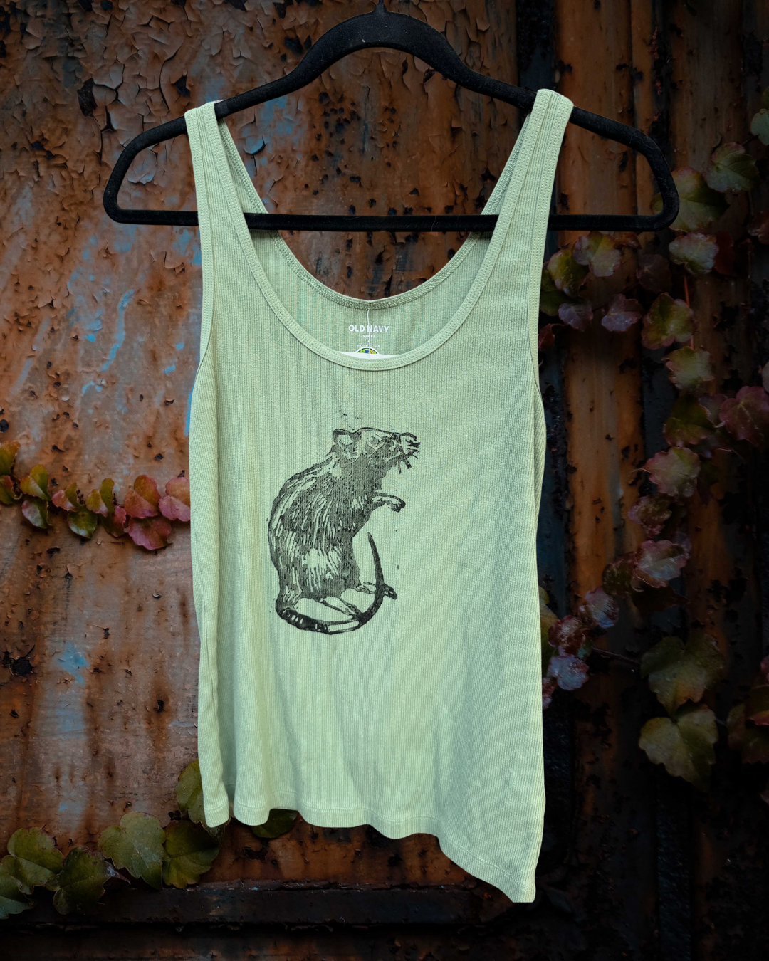 A light sage green tank top with a black rat graphic on the front, hanging on a black hanger against a rustic, weathered brown metal wall with some green leaves.
