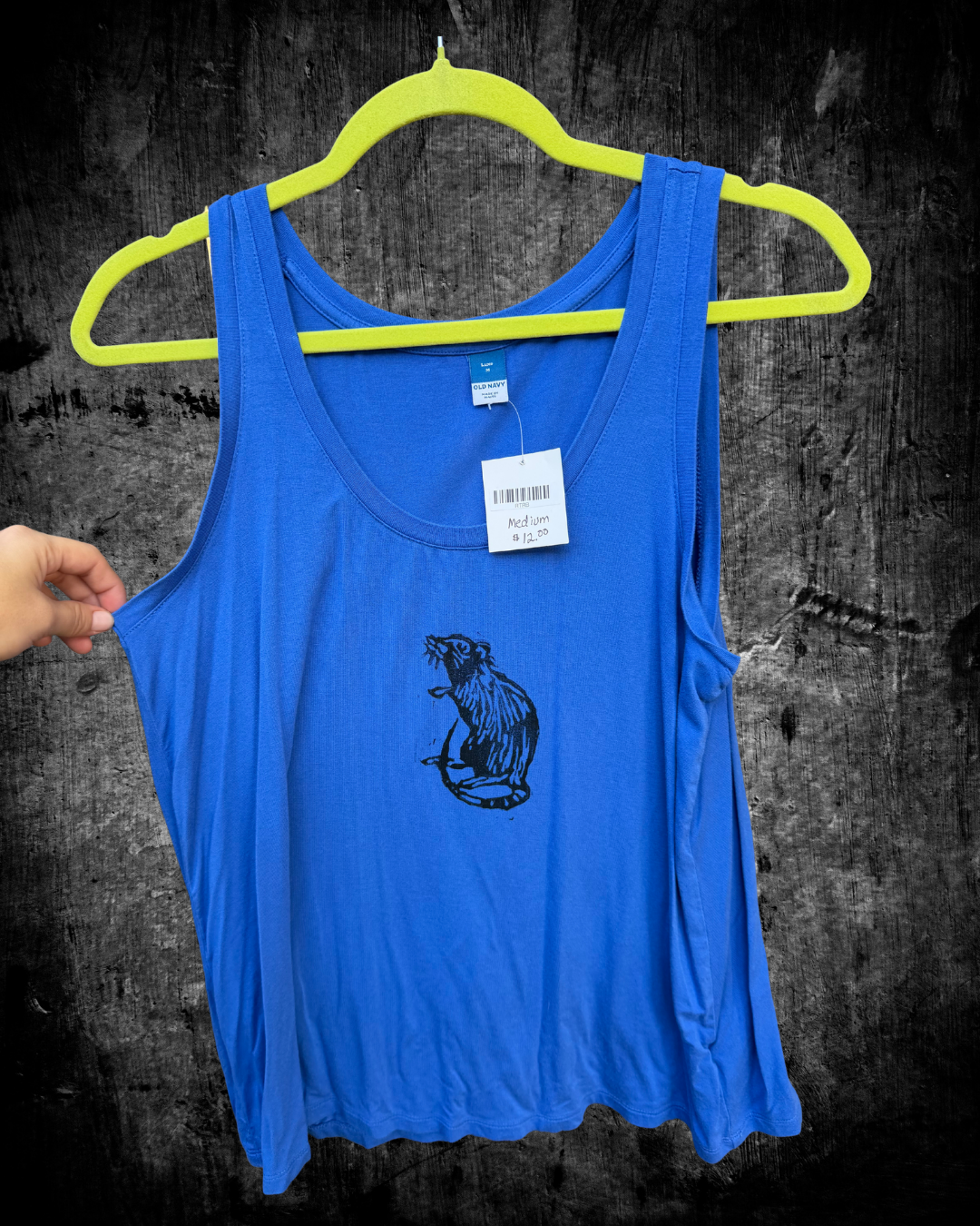 A bright blue sleeveless tank top hanging on a neon yellow hanger against a dark textured wall. A hand is visible on the left holding the tank top out, and a small black graphic of a rat is printed on the front.