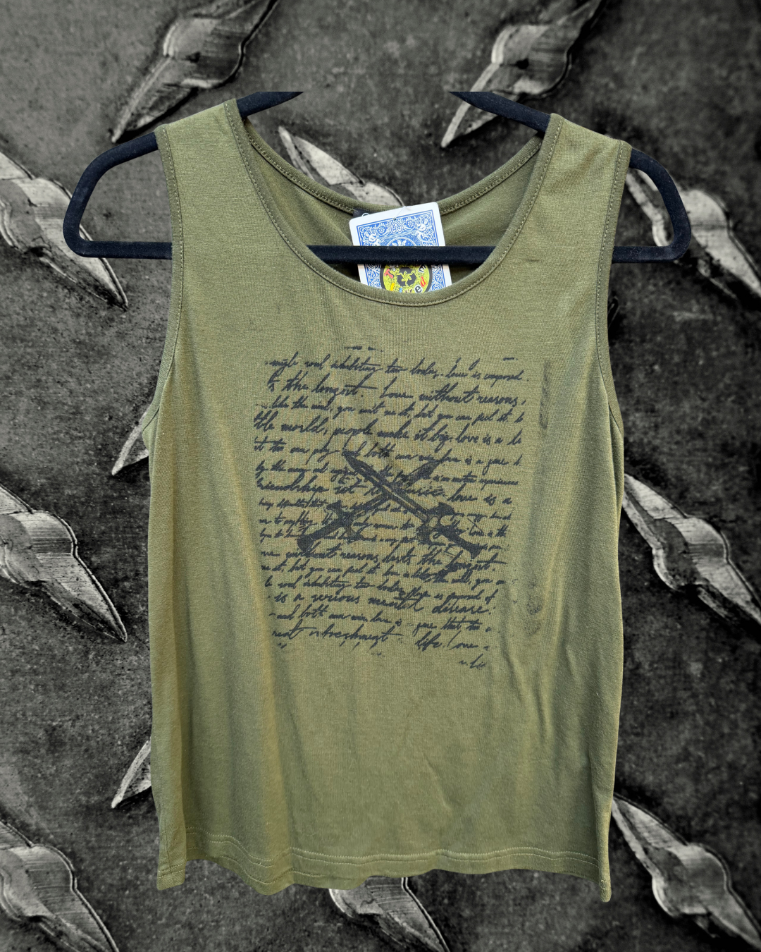 An olive green sleeveless tank top with a black cursive script print and a black graphic sword design on the front, hanging on a black hanger against a dark metal background.