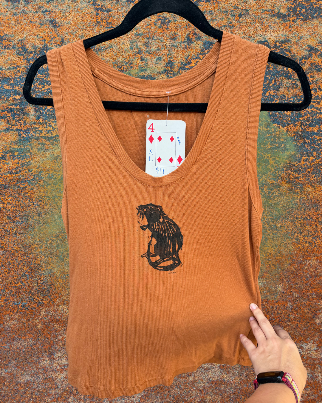 An orange tank top on a black hanger, featuring a black graphic of a rat on the front. A playing card hangtag is attached near the neckline indicating the size is XL, and a hand is holding the bottom right hem of the shirt against a textured, mottled background.